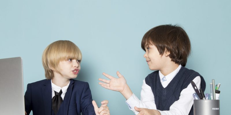 a front view cute business boy in blue classic suit posing in front of silver laptop along with other boy discussing working business work fashion on the blue background